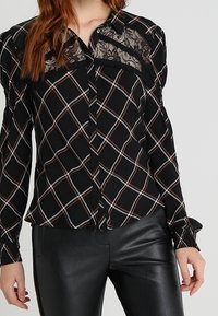 Blusa a quadri nera con yoke in pizzo, dettagli bianchi e marroni, maniche lunghe, colletto classico e chiusura con bottoni.