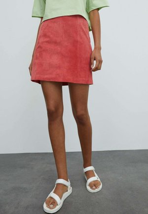 Kvinde iført en koralfarvet ruskind mini nederdel, lysegrøn croppet top og hvide åbne sandaler, stående på en grå gulv mod en ensfarvet baggrund.