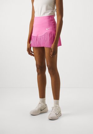 Femme portant une jupe de sport plissée rose et des chaussures de sport blanches avec des chaussettes, debout sur un fond blanc uni.