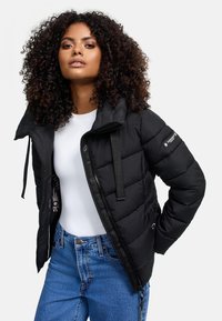 Doudoune noire avec fermeture éclair à l'avant, design rembourré, bretelles ajustables et poches latérales, portée sur un haut blanc et un jean bleu.