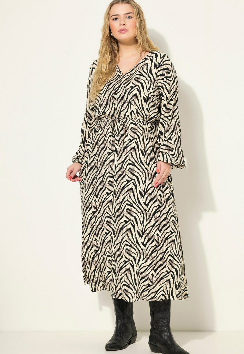 Beige en zwarte maxi-jurk met zebrapatroon, lange mouwen, V-hals en een verstelbare taille. Draagt met zwarte kniehoogte laarzen.