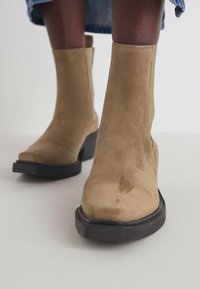 Béžové semišové kotníkové boty s hladkým povrchem, kulatou špičkou a kontrastní silnou černou podrážkou. Elegantní design s minimálními detaily.