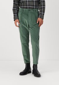 Groene corduroy broek met een taps toelopende vorm, met een textuur op het oppervlak en een enkele plooi aan de voorkant, gecombineerd met zwarte enkellaarzen.