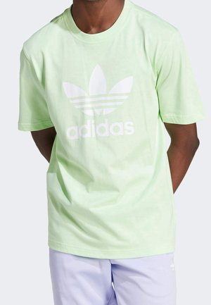 Homme portant un t-shirt Adidas vert clair avec logo tréfle blanc et un short violet clair sur fond uni.