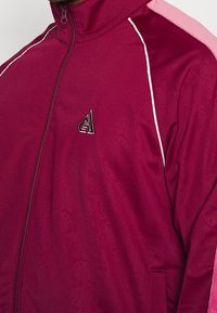 Mörkröd zip-up jacka med ljusrosa detaljer och vit kant. Har en liten logotyp på bröstet och en strukturerad tygfinish.