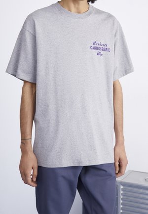 Henkilö, joka on pukeutunut löysään vaaleanharmaaseen t-paitaan, jossa on violetti "Carhartt Carrosserie Wip" -teksti rinnassa, ja tummansiniset housut, seisoo sisätiloissa.