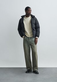 Juoda pūkuota striukė su gobtuvu, dėvima virš kreminės spalvos megztinio. Pora su laisvomis, šviesiai žaliomis džinsomis ir juodais batais. Paprastas fonas.