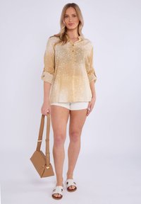 Camicia beige con bottoni e motivo a stelle, maniche arrotolate, abbinata a pantaloni corti bianchi e una borsa marrone. I sandali sono bianchi con design a taglio.