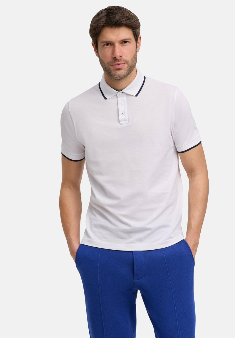 Polo shirt bianco a maniche corte con colletto nero e chiusura a tre bottoni, abbinato a pantaloni blu con texture. Look pulito e informale.