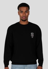 Sudadera negra con un pequeño diseño bordado de una muñeca en el lado izquierdo del pecho, puños acanalados y un cuello redondo clásico.