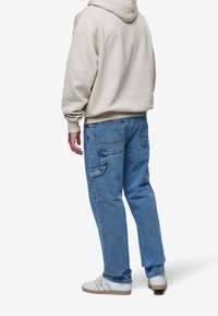 Sudadera con capucha beige, jeans de mezclilla azul con dos bolsillos traseros, parche de marca y zapatillas blancas con acentos grises.
