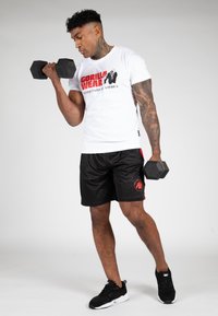 Witte T-shirt met rood "GORILLA WEAR" logo; zwarte shorts met rode accenten; houdt hexagonale dumbbells vast; zwarte sportschoenen.