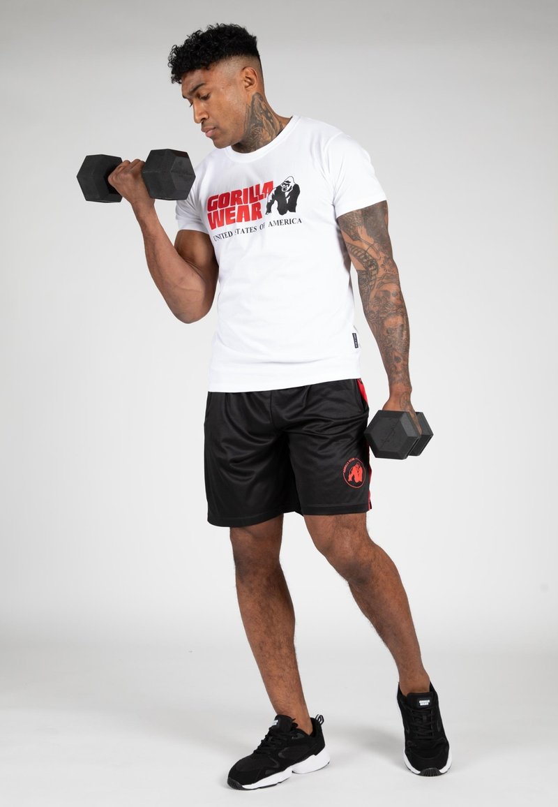 Witte T-shirt met rood "GORILLA WEAR" logo; zwarte shorts met rode accenten; houdt hexagonale dumbbells vast; zwarte sportschoenen.