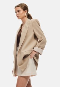 Blazer beige surdimensionné avec des revers en dents de scie, présentant des détails de poignets rayés et des poches latérales. Porté sur un haut marron et un short clair.
