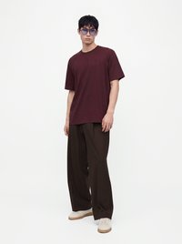 Jeune homme portant un t-shirt rouge foncé, un pantalon large foncé, des baskets blanches et des lunettes de soleil teintées bleu, debout devant un fond clair uni.