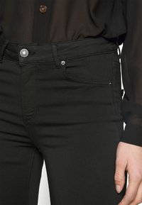 Jeans negros de corte ajustado, con bolsillos frontales, cierre de botón metálico y sutiles detalles de costura a lo largo de la cinturilla.