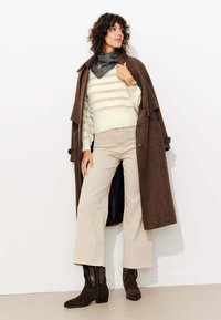 Manteau en laine marron porté sur un pull rayé crème et un pantalon beige. Bottes en daim marron foncé avec un bout pointu et des coutures décoratives.