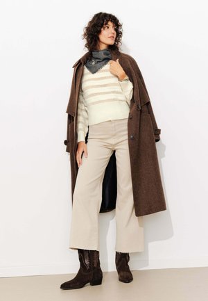 Manteau en laine marron porté sur un pull rayé crème et un pantalon beige. Bottes en daim marron foncé avec un bout pointu et des coutures décoratives.