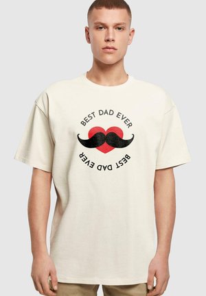T-shirt en coton couleur crème avec un graphique de moustache noire et un cœur rouge, portant l'inscription "MEILLEUR PAPA DE TOUS LES TEMPS" dans un design circulaire.