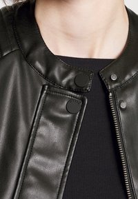 Chaqueta de cuero negro con cuello redondo, botones a presión y cierre de cremallera metálico. Presenta una textura suave y un diseño estructurado.