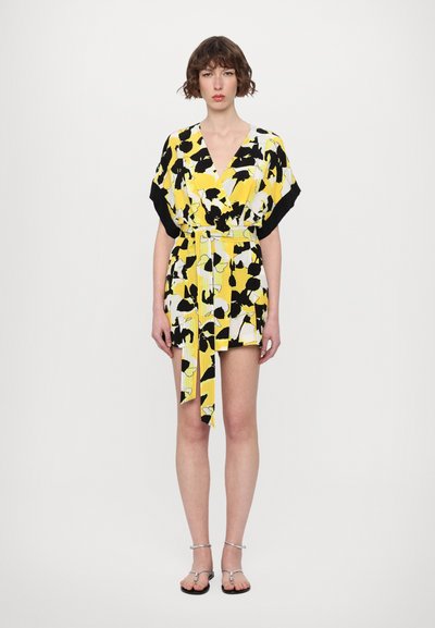 Short, wrap-style dress in yellow with black and white abstract patterns, featuring short black sleeves and a tied belt. Flat sandals.