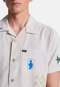 Hombre vestido con camisa beige clara abotonada con diseño abstracto azul en el bolsillo y estrella verde bordada en la manga.