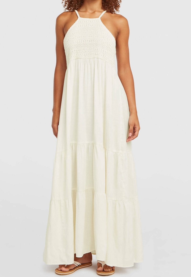 Femme portant une robe maxi sans manches couleur blanc cassé avec un corsage en crochet et une jupe à volants, associée à des sandales plates.