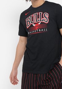 Camiseta de algodón negra con gráfico de "BULLS BASKETBALL" en rojo y blanco, con el logo de Nike en la parte superior izquierda. Mangas cortas, corte regular.