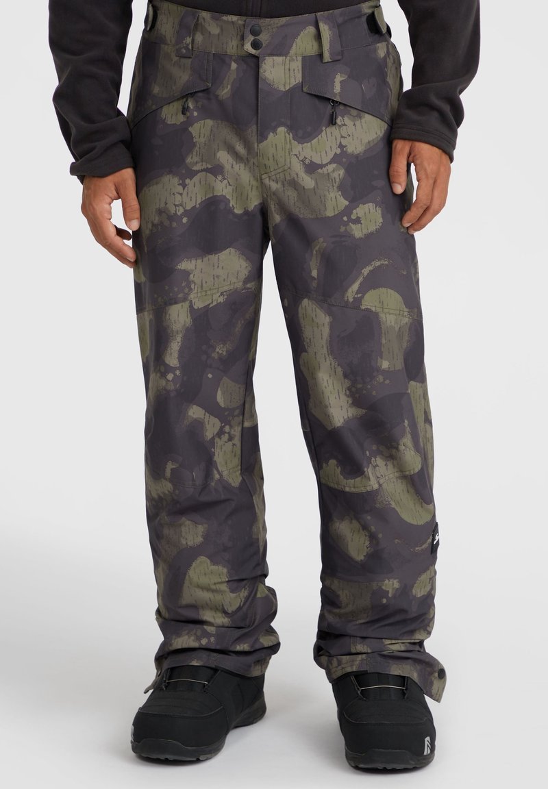 Homme portant un pantalon camouflage noir et vert olive et des bottes d'hiver noires, debout devant un fond clair uni.
