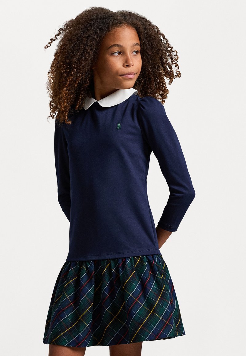 Tamsiai mėlyna suknelė ilgomis rankovėmis su balta apykakle, turinti klostuotą pledo sijoną žalia, raudona ir geltona spalvomis. Apima mažą logotipą ant krūtinės.