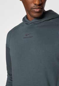 Nike Pro mikina v tmavosivej tkanine, s prednou kapsou, žebrovanými manžetami a malým potlačeným logom na hrudi. Hladký povrch.