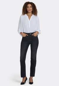 Vit blus med långa ärmar och knappslå, kombinerad med höga svarta jeans och svarta klackskor, presenterad mot en neutral bakgrund.