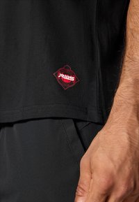 Sort t-shirt med et rødt diamantformet logo, der har "RWS" og "Stadium". Glat bomuldsstof, korte ærmer og en lige skæredesign.