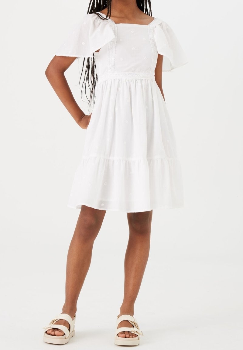 Jeune femme portant une robe blanche aux épaules dénudées, longueur genou, avec des manches bouffantes et des sandales blanches, debout les mains sur les hanches.