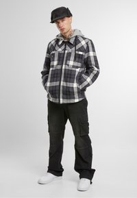 Svart og hvitt rutet flanelljakke med en grå hettegenser, to frontlommer, kombinert med svarte cargo-bukser og hvite sneakers. Svart caps på hodet.