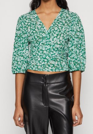 Femme portant un chemisier vert style cache-cœur avec un imprimé floral blanc et un pantalon noir taille haute en similicuir.