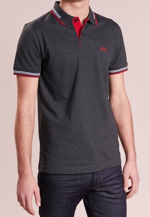 Mann trägt ein dunkelgraues Polohemd mit rot-weiß gestreiftem Kragen und Ärmelabschlüssen, kombiniert mit dunkelblauen Jeans.