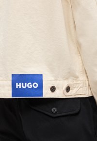 Μπεζ στρίφωμα μπουφάν με μπλε ετικέτα "HUGO" και μεταλλικά κουμπιά πάνω από μαύρο παντελόνι με πατιλέτα τσέπης με κουμπί.