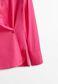 Lyserød skjorte med knapper, lange ærmer, lige snit, sideslids og blød tekstur. Knapperne er i en mørkere lyserød nuance.