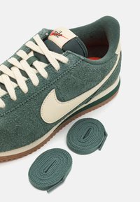 Zelené semišové tenisky Nike s krémovými tkaničkami, krémovým symbolem Nike a vedle nich dvěma extra zelenými tkaničkami na bílém pozadí.