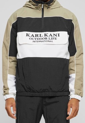 Homme portant une veste à capuche à blocs de couleurs noir, blanc et beige avec une poche avant à rabat et le texte « KARL KANI OUTDOOR LIFE INTERNATIONAL ».