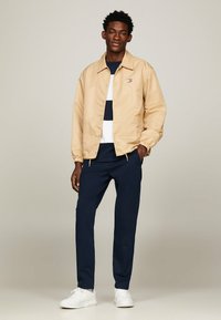 Giacca leggera beige con polsini elastici, pantaloni blu navy e sneakers bianche. La parte superiore ha un motivo a righe in blu navy e bianco.