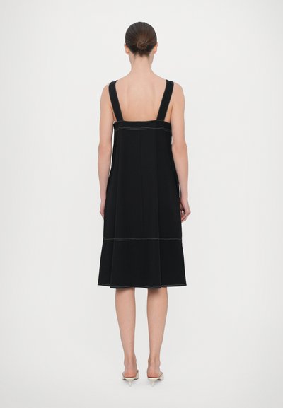 Crna haljina bez rukava s širokim naramenicama, otvorenim leđima i kontrastnim šavovima. Lepršavi A-linijski kroj završava tik ispod koljena.