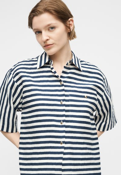 Γυναίκα με κοντά ανοιχτό καστανά μαλλιά που φοράει ένα μπλε ναυτικό και λευκό πουκάμισο με οριζόντιες ρίγες, κοντά μανίκια, κουμπιά και γιακά.