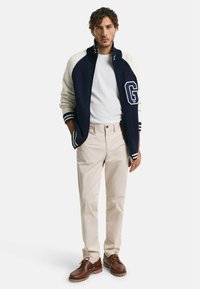 Chaqueta estilo varsity en azul marino y crema con rayas blancas, camiseta blanca, pantalones beige claros y zapatos marrones con cordones y suela robusta.
