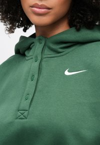 Sweat à capuche vert en tissu doux avec une patte de boutonnage. Présente un logo Nike blanc sur la poitrine et une capuche ajustable avec cordon de serrage.