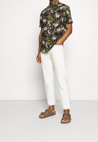 Lyhythihainen musta paita kirkkaalla kukkakuviolla, yhdistetty valkoisiin kapealahkeisiin housuihin ja ruskeisiin sandaaleihin, joissa on kaksi säädettävää remmiä.