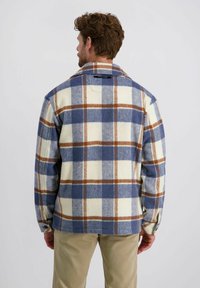 Man met zijn rug naar de camera gekeerd, gekleed in een blauw, wit en bruin geruit jasje met beige broek tegen een effen achtergrond.