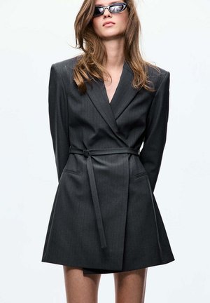 Mujer vistiendo un vestido tipo blazer gris oscuro de gran tamaño con cinturón y gafas de sol negras, posando frente a un fondo blanco liso.