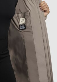 Chaqueta ligera beige abierta para mostrar el interior, con textura acolchada, dos bolsillos internos y etiquetas de marca cosidas en el interior.